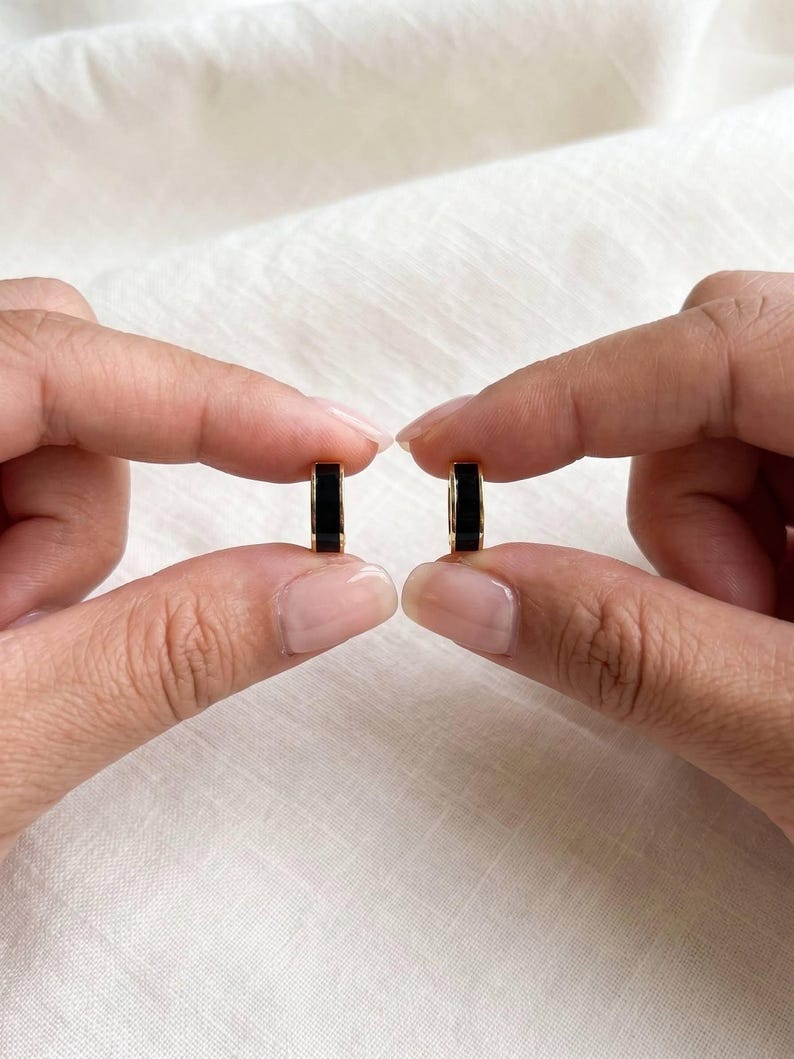 Two hands holding black enamel huggie hoop earrings