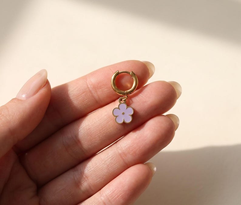 Hand holding gold huggie hoop earring with purple enamel flower charm